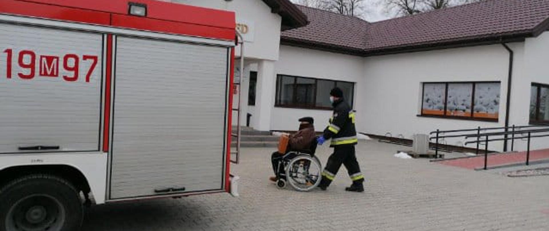 Zdjęcie przedstawia strażaka, który przewozi seniora na wózku inwalidzkim. 