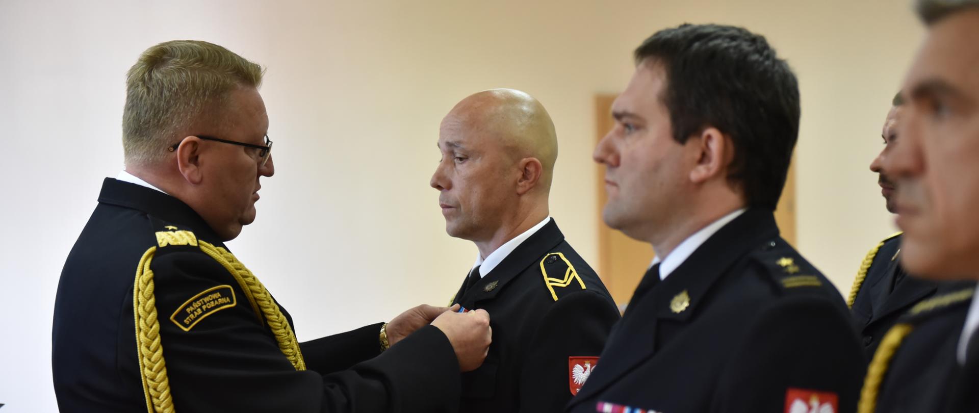 Zdjęcie przedstawia strażaka Państwowej Straży Pożarnej w stopniu nadbrygadiera, który odznacza innych strażaków odznaczeniami.