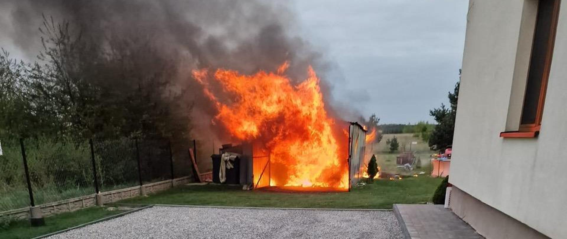 Zdjęcie przedstawia prywatną posesję, na której pali się wnętrze wolnostojącego garażu typu „blaszak”. Po prawej stronie Widać dom mieszkalny.