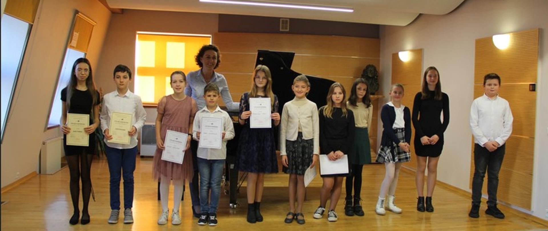 Zdjęcie przedstawia stojące na scenie dzieci, uczestników konkursu pianistycznego wraz z panią wicedyrektor.