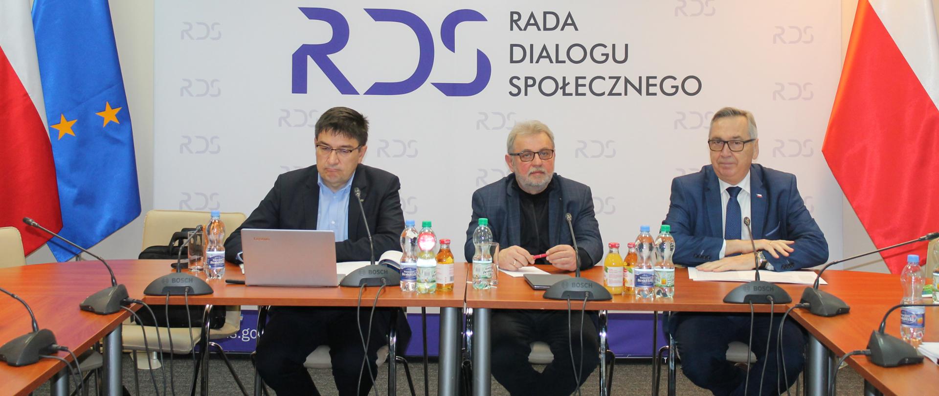 Na zdjęciu widnieje stół prezydialny za którym siedzą od lewej strony: Wojciech Ilnicki - Przewodniczący zespołu ds. rozwoju dialogu społecznego, Henryk Nakonieczny - Przewodniczący zespołu ds. prawa pracy oraz Stanisław Szwed - Sekretarz Stanu w Ministerstwie Rodziny i Polityki Społecznej. Po lewej stronie widnieją dwie flagi: Polski i UE a po prawej stronie flaga Polski. W tle siedzących znajduje się baner z napisem Rada Dialogu Społecznego a nad nim wisi godło Polski.