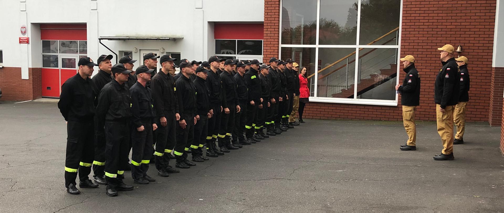 Strażaków stojących w dwuszeregu podczas zbiórki. Naprzeciwko nich stoi komendant, jego zastępca oraz dowódca JRG. Komendant przemawia do strażaków.