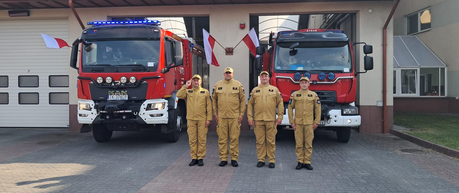 4 strażaków na tle garażu i 2 wozów strażackich , na dole kostka brukowa 