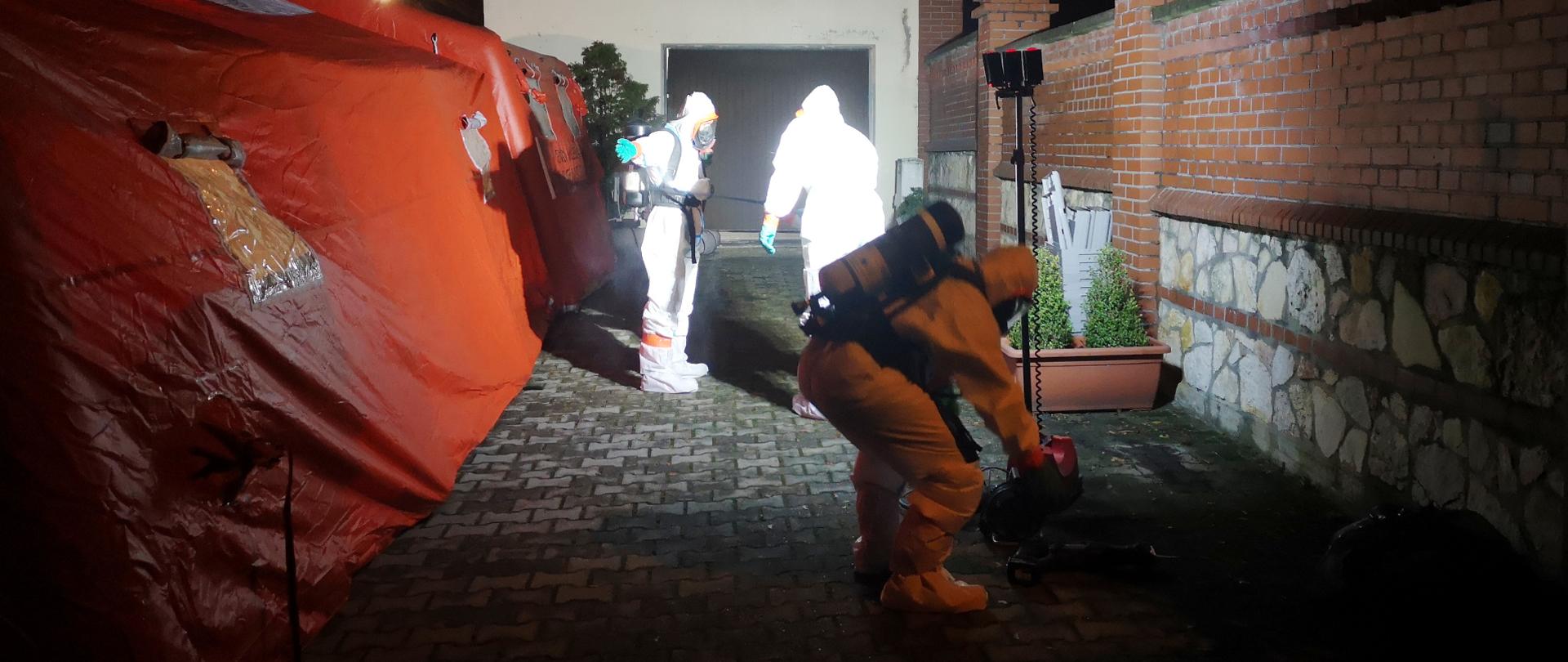 Na zdjęciu widać 3 strażaków w lekkich kombinezonach przeznaczonych do ochrony przed zagrożeniami chemicznymi i biologicznymi, którzy przygotowują się do wstępnej dekontaminacji. Oświetleni są reflektorem. Stoją obok dwóch namiotów pneumatycznych w kolorze pomarańczowym.