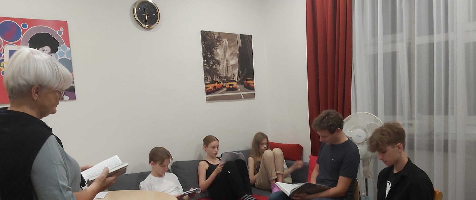 Po lewej stronie na pierwszym planie stoi pani Marzena, inicjatorka akcji. Na kanapie siedzi trzech wychowanków, mają przed sobą teksty powieści. Po prawej stronie są 2 osoby z tekstami powieści.