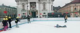 Widok z boku. Uczestnicy pokazu - dzieci z dziecięcej drużyny pożarniczej stoją na lodowisku przy lewej bandzie. Po prawej stronie dziecko symulujące zarwanie się pod nim tafli lodu. Trzyma rzutkę z liną, pozostali ciągną linę i symulują wydostawanie osoby na brzeg. Na środku opiekun MDP. Wokół bandy widownia. W tle bazylika.