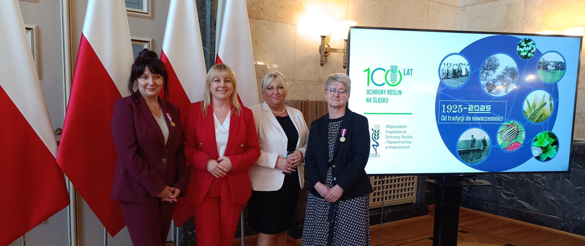 Trzy kobiety ubrane na elegancko. Stoją a za nimi są biało-czerwone flagi. Po prawej stronie stoi duży monitor z wyświetlonym obrazem.