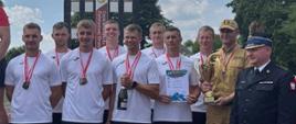 Strażacy w białych sportowych koszulkach na 3 miejscu podium z medalami i pucharem. 