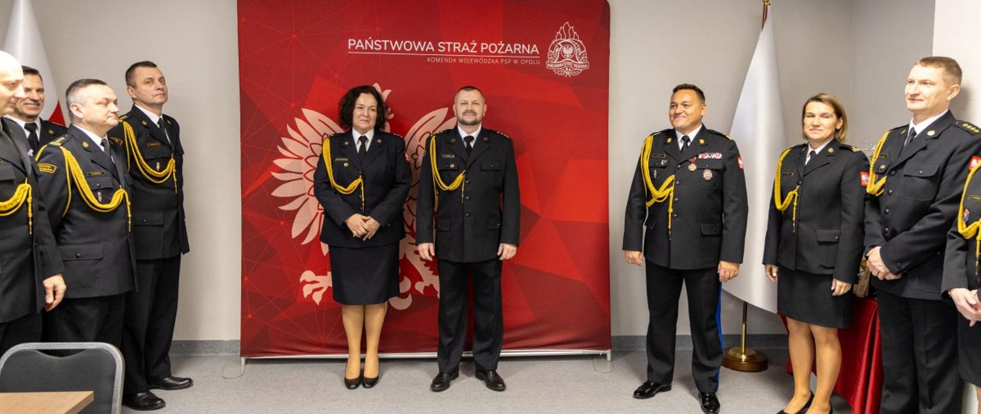 Pożegnanie naczelnik wydziału kadr KW PSP Opole oraz zastępcy naczelnika Ośrodka Szkolenia KW PSP Opole. Na zdjęciu widać Pani naczelnik, Pana zastępcę naczelnika, Opolskiego Komendanta Wojewódzkiego PSP wraz z zastępcami oraz komendantów powiatowych / miejskich PSP i pracowników KW PSP Opole w umundurowaniu wyjściowym. W tle Godło RP na czerwonym tle.