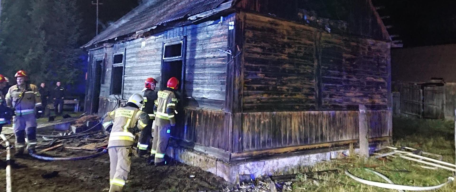 Na pierwszym planie drewniany budynek mieszkalny okopcony bez okien. Przed budynkiem znajdują się strażacy oraz 2 policjantów. Wokół domu nadpalone deski i elementy konstrukcji. 