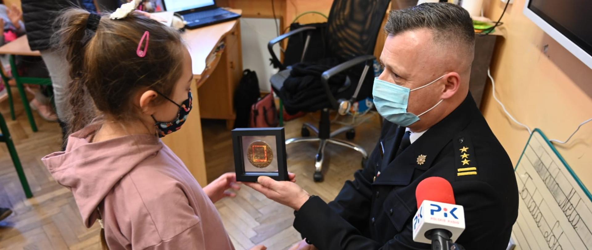 Zdjęcie przedstawia strażaka w galowym ciemnym mundurze w pozycji na kucka wręczającego odznaczenie małej dziewczynce w pomieszczeniu klasowym. Oboje mają założone maseczki ochronne. W tle wyposażenie klasy.