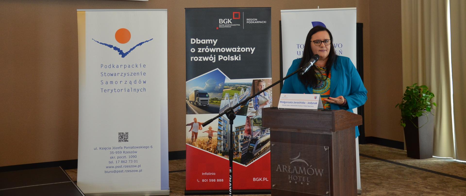 Wiceminister Małgorzata Jarosińska-Jedynak w mównicy z mikrofonem na tle banerów