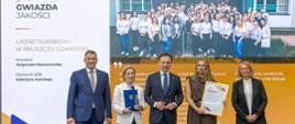 Pięć osób stoi na tle baneru Gwiazda Jakości. Dwie kobiety trzymają w ręku dyplomy.
