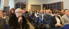 Na sali konferencyjnej w kilku rzędach siedzą uczestnicy spotkania i słuchają osoby mówiącej do mikrofonu