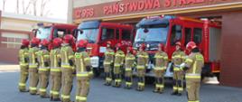 Zdjęcie przedstawia strażaków na zmianie służby przed budynkiem Komendy Powiatowej na tle samochodów pożarniczych oddających hołd poległy strażakom z Ukrainy 
