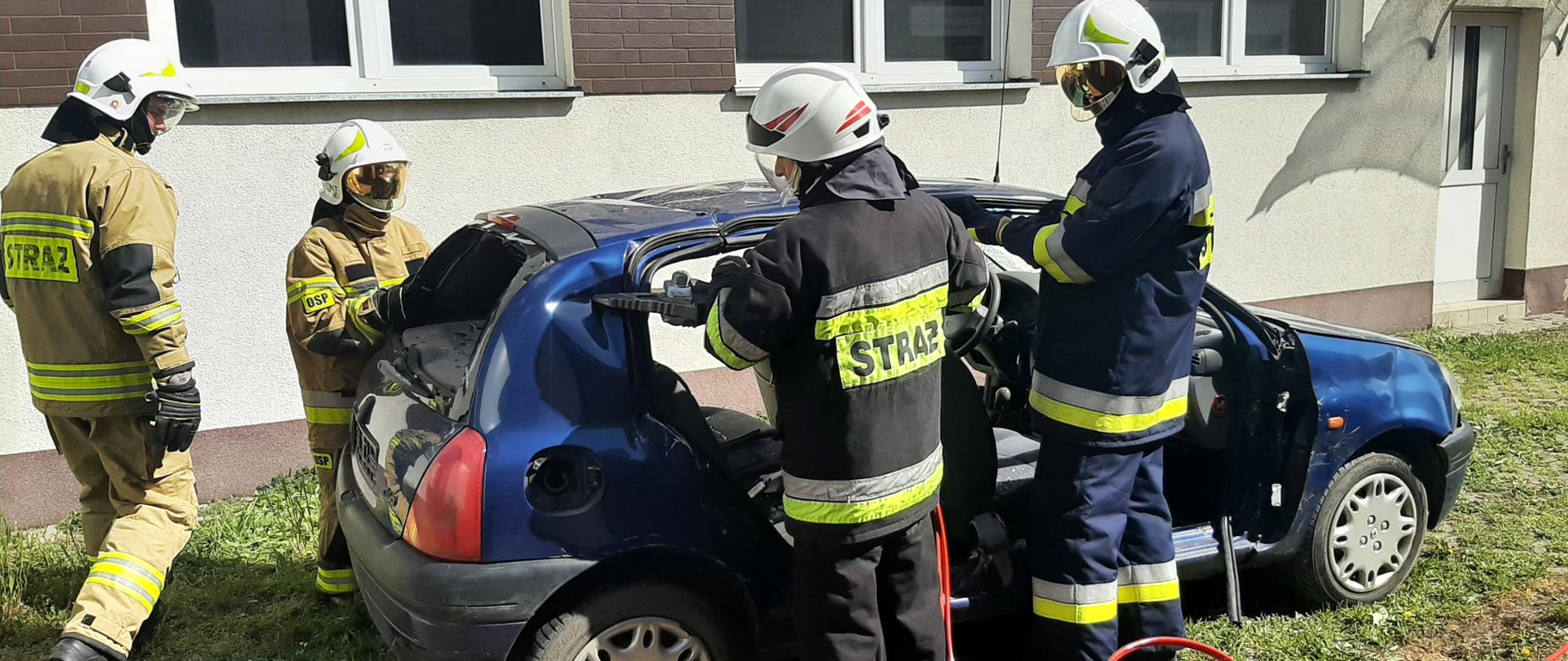 Zdjęcie przedstawia czterech strażaków ubranych w hełmy i ubrania specjalne rozcinających karoserię samochodu osobowego przy pomocy nożyc hydraulicznych. Auto znajduje się na betonowej ażurowej płycie z której wyrasta trawa, w tle widnieje ściana jasnego budynku z oknami.