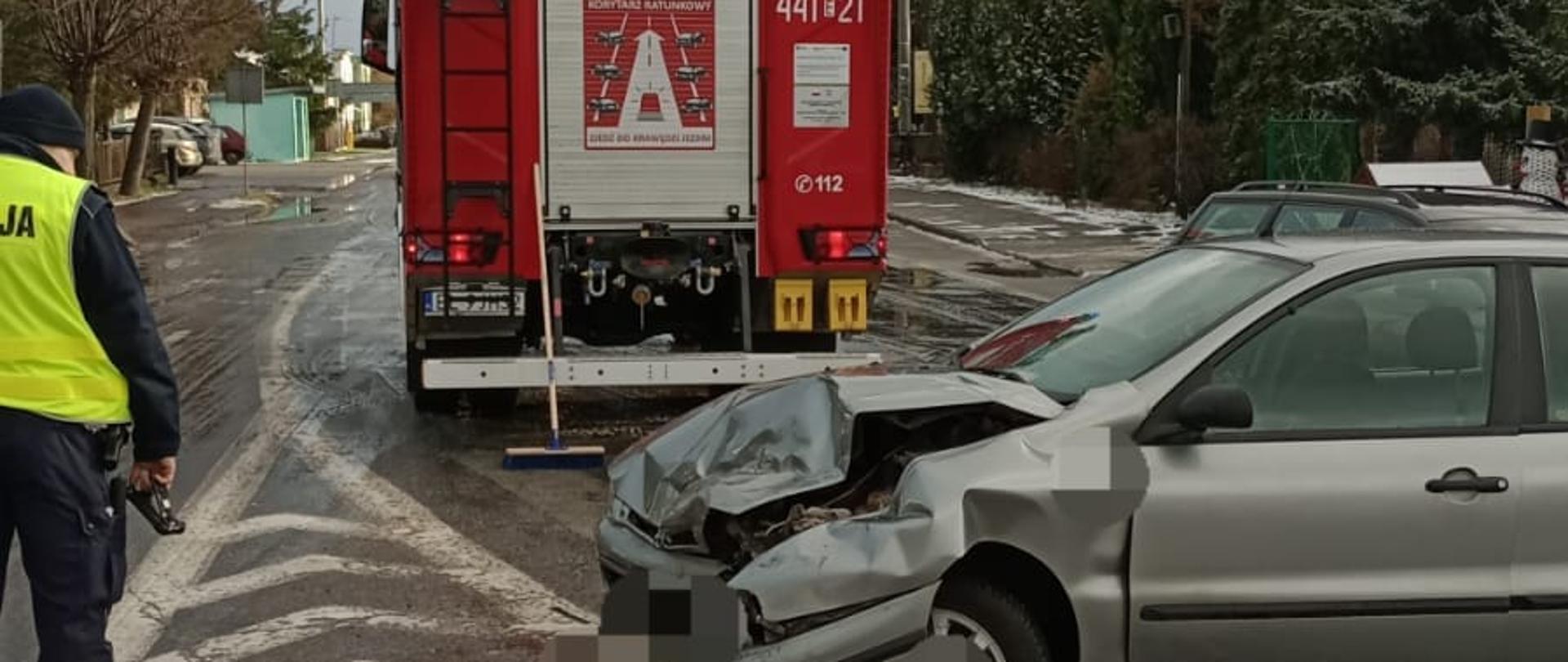 Na zdjęciu widać rozbity srebrny samochód osobowy stojący na środku asfaltowej jezdni. Z tyłu stoi czerwony duży samochód strażacki. Obok stoi policjant. W tle widać inne samochody oraz drzewa. Jest widno, ale pochmurnie.