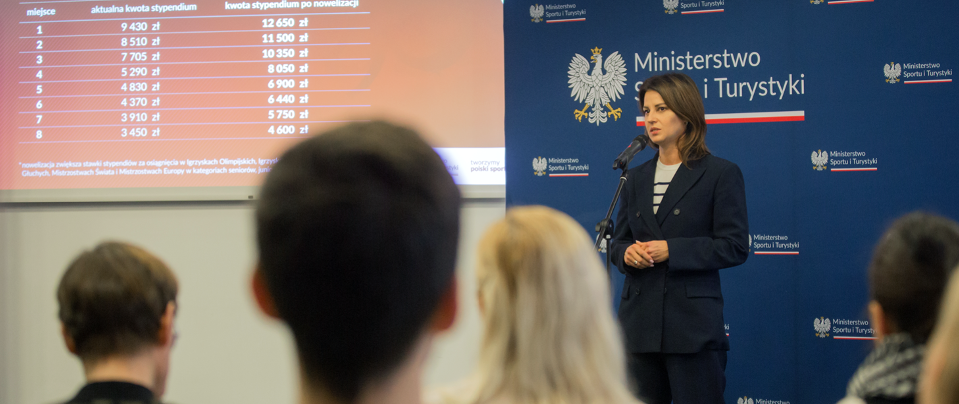 The Minister of Sport and Tourism speaking in front of the crowd, the backgrond slide shows the amount of money for athletes