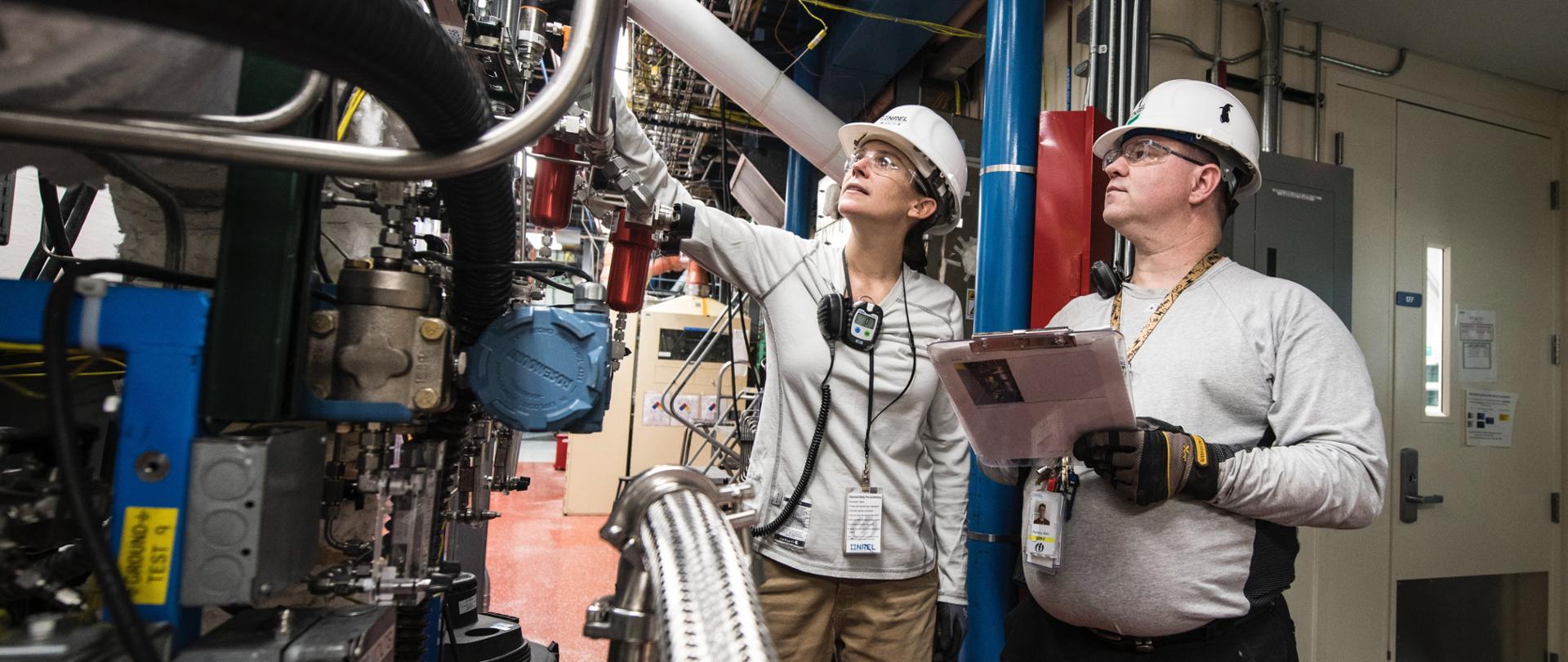 Ludzie w kaskach, za nimi przemysłowe instalacje.