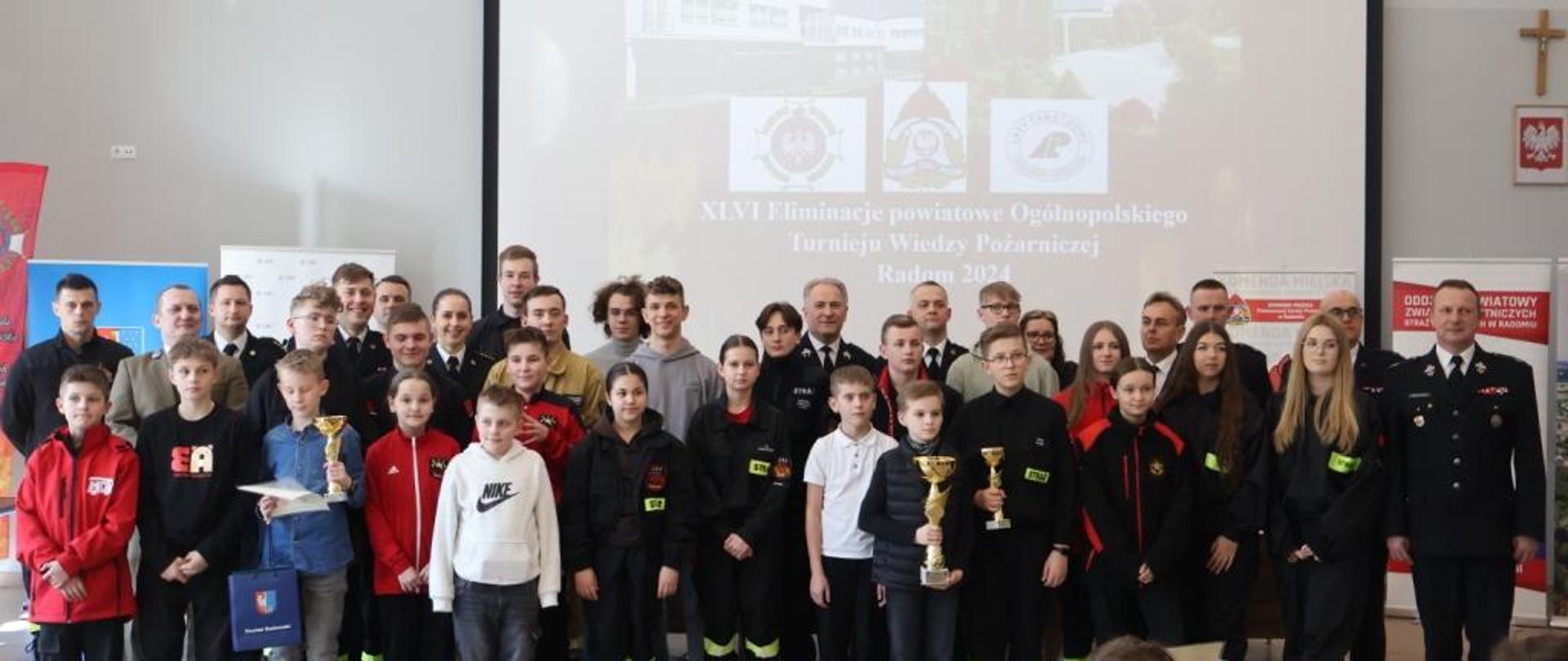 Pamiątkowe zdjęcie wykonane na zakończenie turnieju. Na zdjęciu widać wszystkich uczestników turnieju. Niektórzy z nich jako zwycięzcy w dłoniach trzymają pamiątkowe puchary, dyplomy oraz upominki. Za nimi w rzędzie stoją przedstawiciele Lasów Państwowych, funkcjonariusze Państwowej Straży Pożarnej, Prezesi jednostek z Ochotniczych Straży Pożarnych oraz przedstawiciele lokalnych władz samorządowych. 