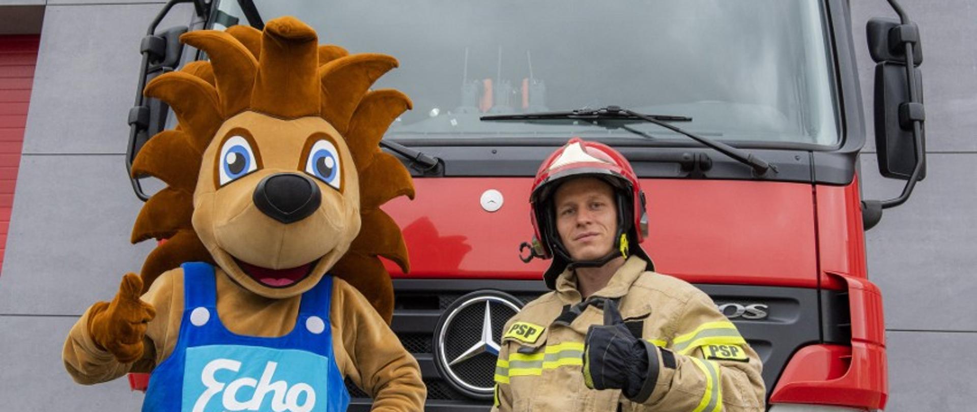Zdjęcie przedstawia strazaka ubranego w ubranie specjalne oraz hełm i maskotkę jeża Edka stojących na tle wozu strażackiego.
