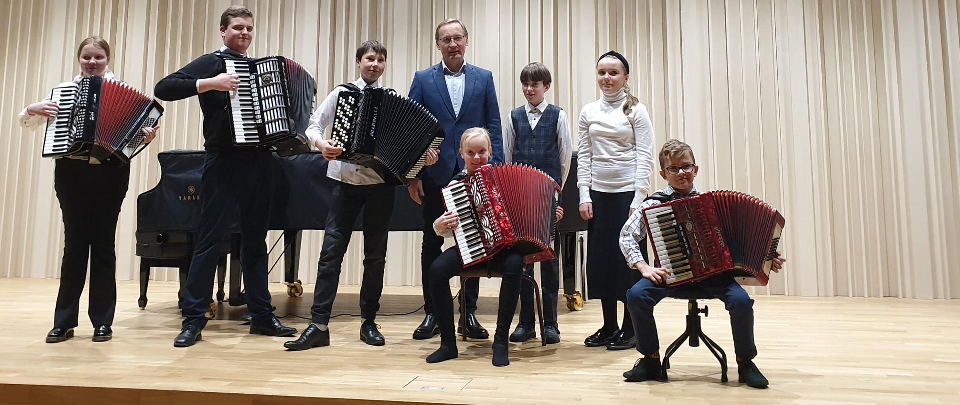 Na zdjęciu grupa uczniów, którzy trzymają akordeony. Za nimi stoi mężczyzna w okularach w granatowym garniturze.