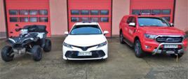 Na tle garażu Komendy widoczne nowo zakupione pojazdy, od prawej: terenowy czerwony Ford Ranger, Biała toyota Camry (samochód operacyjny), oraz quad.