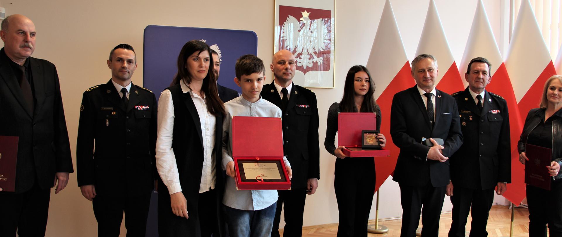 Pamiątkowe zdjęcie grupowe. Odznaczeni odznaką Iuvenis Forti: Zosia Cerkowniak i Patryk Opałko oraz wojewoda świętokrzyski Zbigniew Koniusz, zastępca świętokrzyskiego komendanta wojewódzkiego Państwowej Straży Pożarnej st. bryg. Grzegorz Rajca, komendant miejski PSP w Kielcach st. bryg. Sławomir Karwat, komendant powiatowy st. bryg. Artur Brachowicz, dyrektor II LO im. Jana Śniadeckiego w Kielcach Tomasz Kośny i inni.