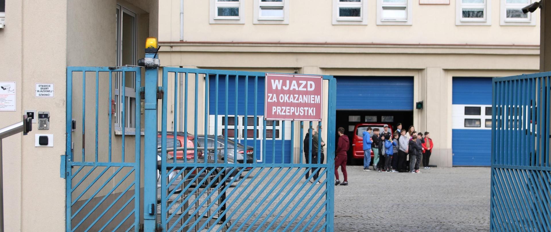 Niebieska brama wjazdowa na plac wewnętrzny SAPSP w Krakowie. Jedno skrzydło uchylone do połowy w tle grupa ludzi. 