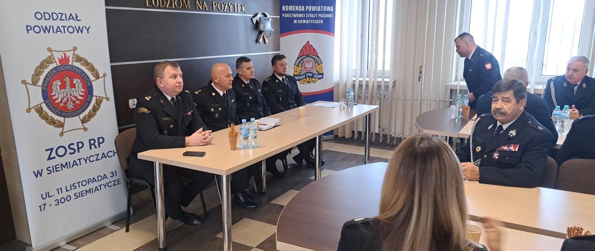 Przy stole siedzi czterech mężczyzn w strażackich mundurach. Obok kilku strażaków, również w strażackich mundurach.
