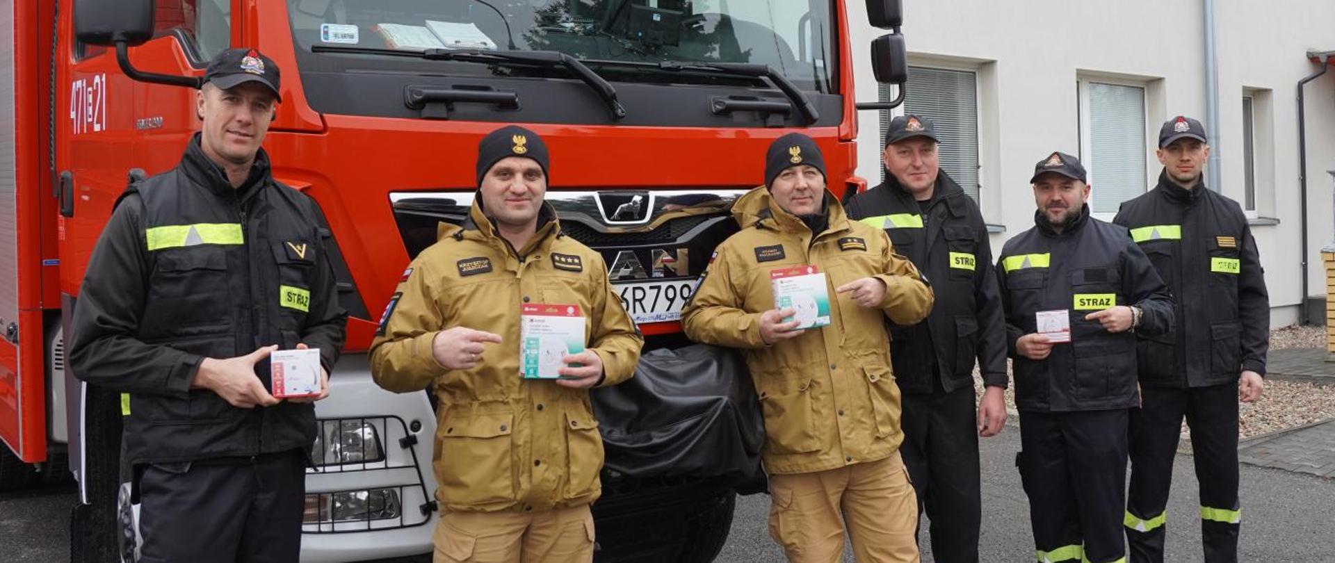 Grupa sześciu strażaków w ubraniach koszarowych stojąca przed samochodem ratowniczo - gaśniczym, trzyma w dłoniach czujki tlenku węgla. Po środku stoją Komendanci Powiatowi i wskazują palcami na czujki tlenku węgla. W tle budynek Komendy Powiatowej Straży Pożarnej w Opocznie. 