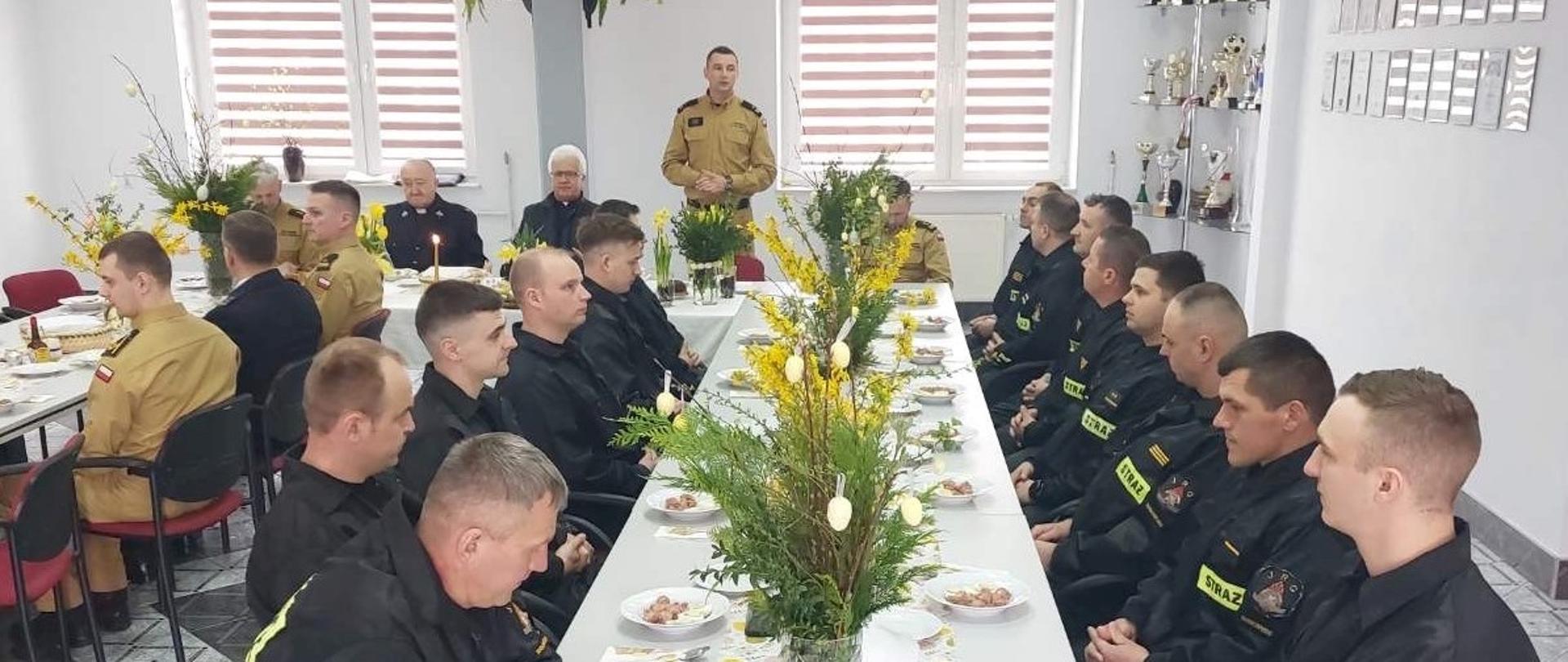 przy świątecznie przyzdobionym stole, w dużym pomieszczeniu siedzą strażacy. Jeden ze strażaków przemawia stojąc. 