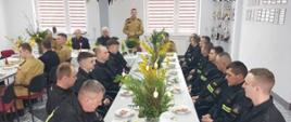 przy świątecznie przyzdobionym stole, w dużym pomieszczeniu siedzą strażacy. Jeden ze strażaków przemawia stojąc. 