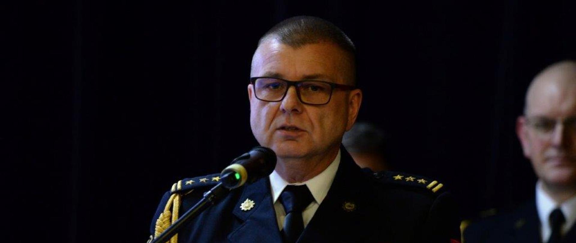 1. Komendant Powiatowy Państwowej Straży Pożarnej w Toruniu stoi przy mównicy z mikrofonem. Komendant ma na sobie strażacki mundur wyjściowy. W tle widać p.o. Kujawsko-Pomorskiego Komendanta Wojewódzkiego. Z tyłu na ścianie znajduje się ciemnogranatowa kotara. 
