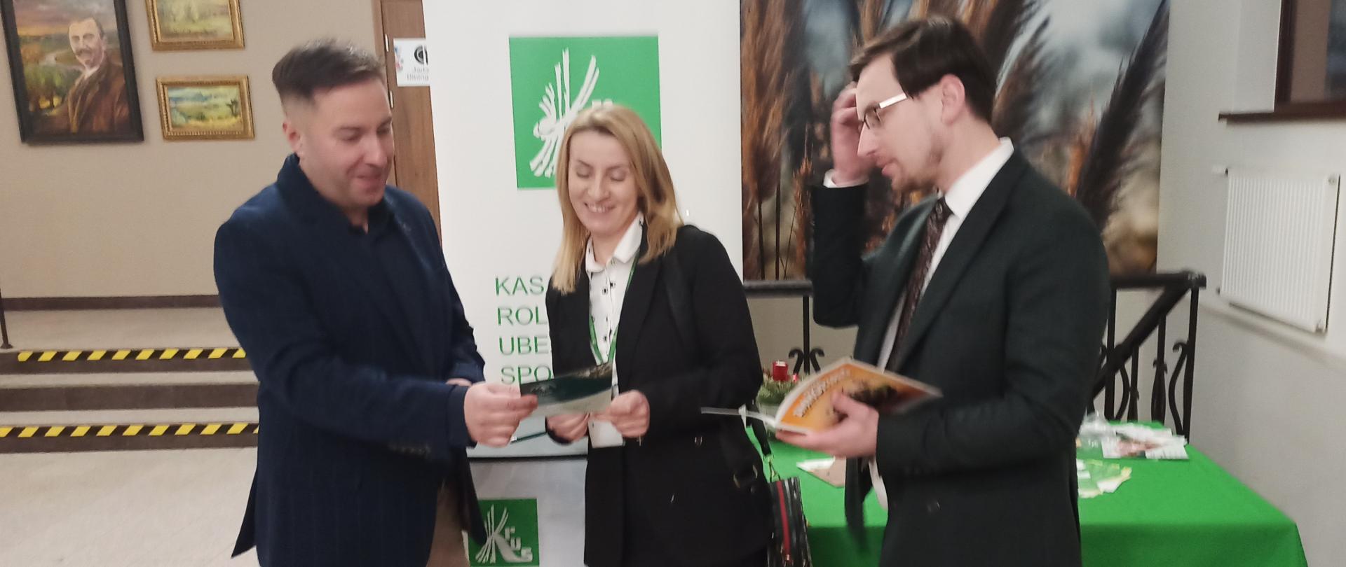 Trzy osoby stoją przy stoisku informacyjnym KRUS. Dwóch mężczyzn i kobieta oglądają materiały informacyjne. Za nimi biało-zielony rollup z logo KRUS i stolik z materiałami informacyjnymi przykryty zielonym obrusem z logo KRUS.