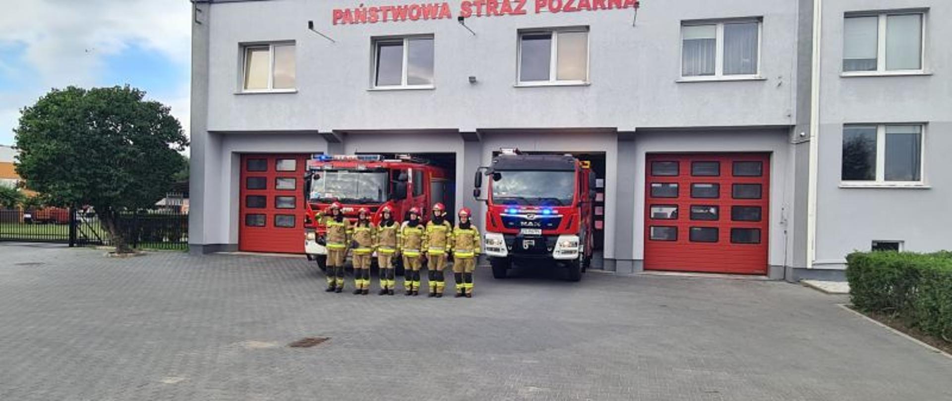 Zdjęcie zrobione na parkingu przed Komendą. Centralny punkt zdjęcia stanowią stojący strażacy. Strażacy stoją w szeregu mają na sobie piaskowe munduru i czerwone hełmy. W tle budynek komendy i czerwone pojazdy gaśnicze.