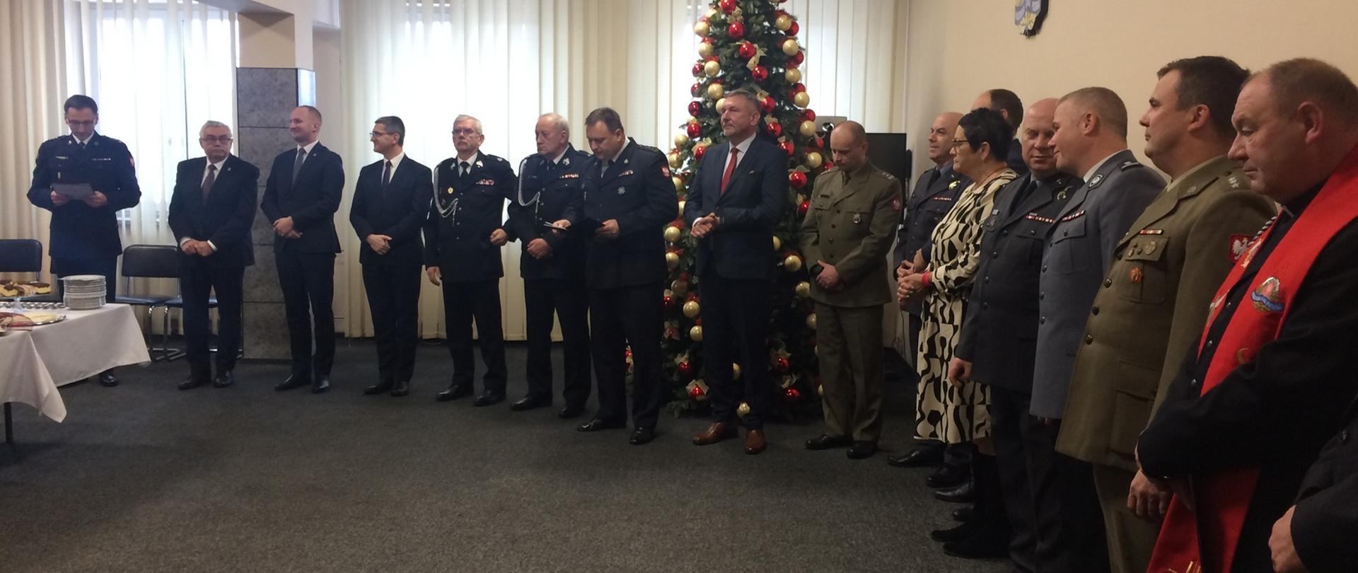 Zdjęcie przedstawia wnętrze sali konferencyjnej w komendzie powiatowej psp w łasku, na tle ściany i choinki stoją zaproszeni na wigilię goście.