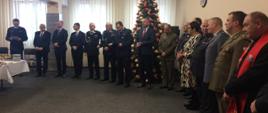 Zdjęcie przedstawia wnętrze sali konferencyjnej w komendzie powiatowej psp w łasku, na tle ściany stoją zaproszeni na wigilię goście, widać również choinkę.