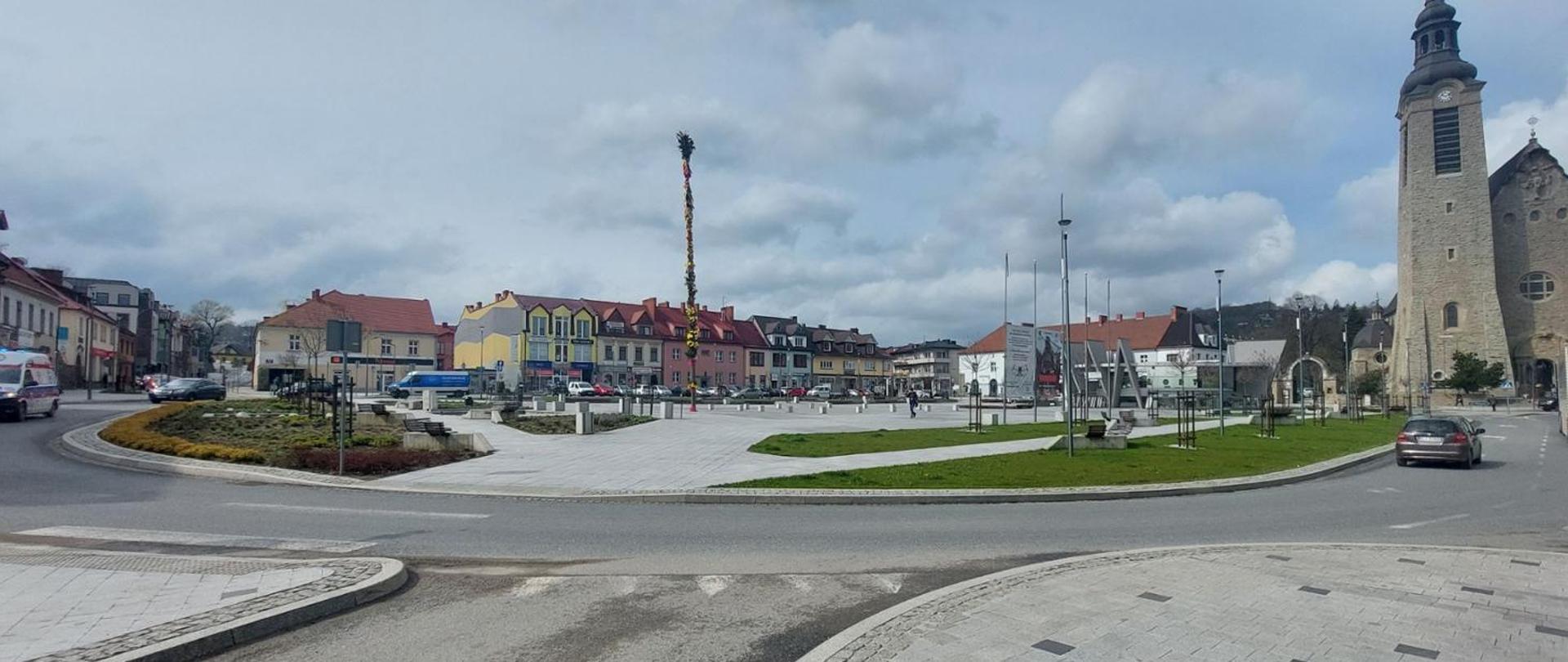 Na zdjęciu widać rozległy rynek w małym mieście, z centralnym placem otoczonym niską zabudową o kolorowych fasadach – w odcieniach żółci, czerwieni i pastelowych barw. Po lewej stronie znajduje się fragment ronda z zielonymi nasadzeniami i krzewami. 