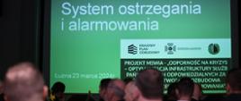 Zajęcie przedstawia strażaków Ochotniczych Straży Pożarnych, podczas narady operacyjnej dla naczelników Ochotniczych Straży Pożarnych i komendantów gminnych ZOSP RP z terenu powiatu gorlickiego. Strażacy OSP ubrani w mundury wyjściowe granatowe i białe koszule strażacy PSP w ubrania służbowe koloru piaskowego. Narada ma miejsce w sali Gminnego Ośrodka Kultury w Łużnej strażacy siedzą przy stołach patrząc na ekran z napisem białym na zielonym tle napis koloru białego "System ostrzegania i alarmowania".