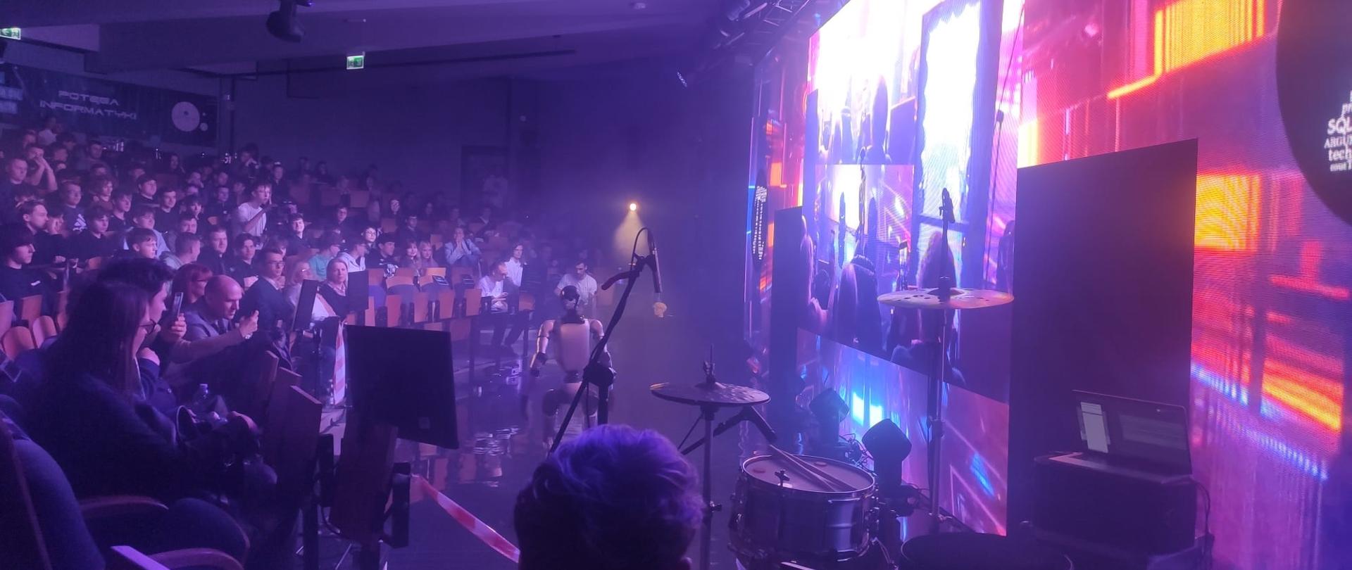 Widok sceny z publicznością, kolorowe światła i elementy techniczne.