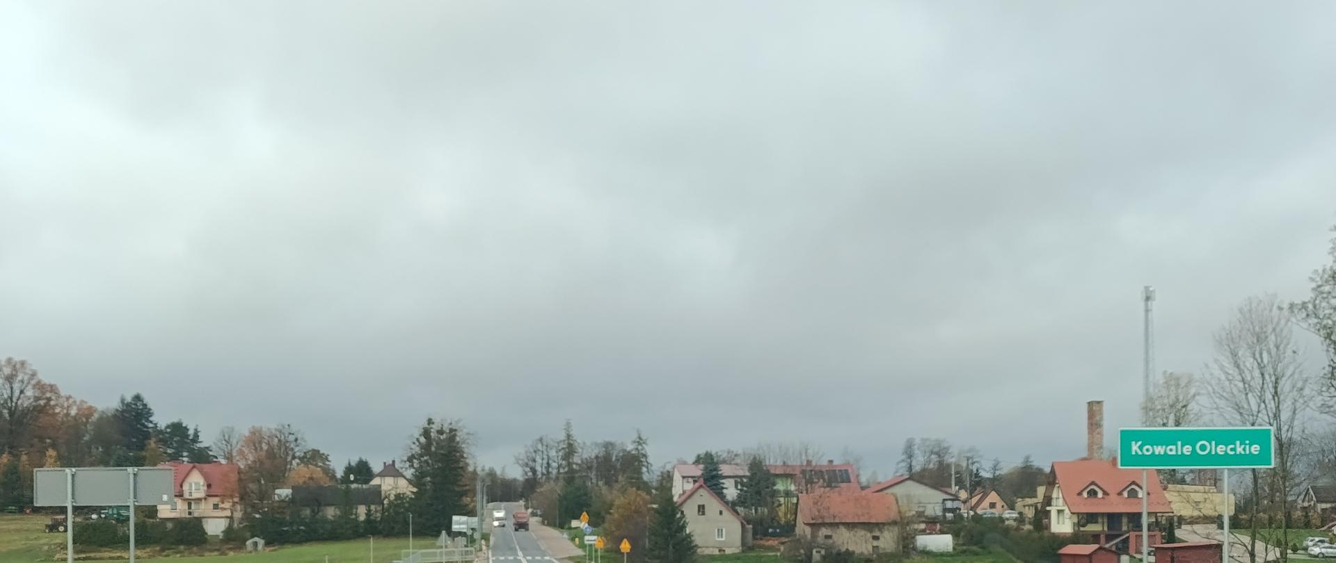 Zdjęcie przedstawia nową, asfaltową drogę z podwójną linią ciągłą na środku, prowadzącą w kierunku niewielkiej miejscowości. Po obu stronach drogi znajdują się metalowe bariery ochronne oraz zielone pola. W tle widać zabudowania wsi z kilkoma domami o jasnych elewacjach i czerwonych dachach, a także komin i maszt telekomunikacyjny. Po prawej stronie drogi stoi zielona tablica z białym napisem „Kowale Oleckie”, wskazująca nazwę miejscowości. Niebo jest pochmurne i szare, co sugeruje jesienną aurę. 