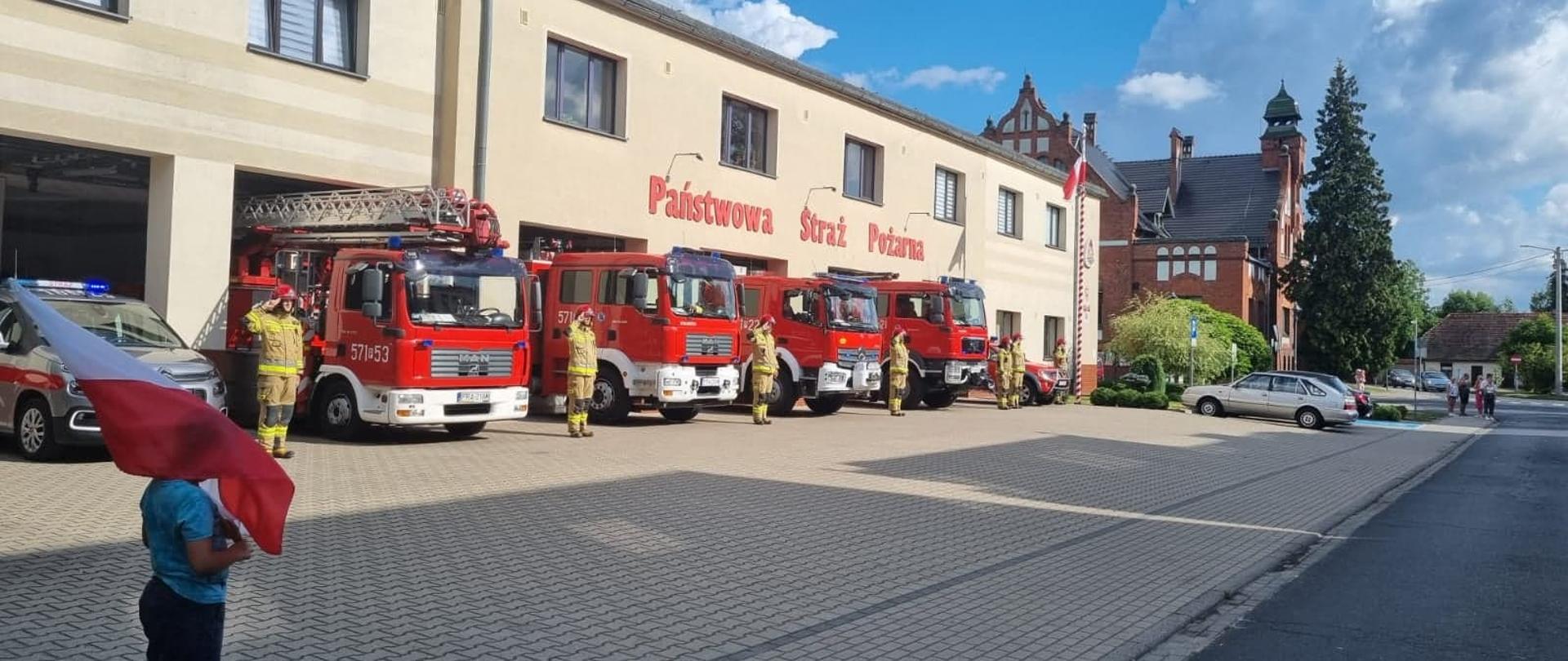 Rocznica wybuchu Powstania Warszawskiego. Plac przed Komendą Powiatową PSP w Rawiczu. Przed garażami stoją wozy bojowe z włączonymi sygnałami świetlnymi. Obok każdego pojazdu stoi strażak w ubraniu specjalnym, który salutuje. Na pierwszym planie dziecko trzymające w rękach biało-czerwoną flagę.