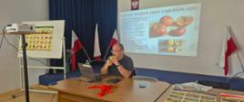 Mężczyzna siedzi przy dużym stole konferencyjnym i trzyma przy ustach jabłko podczas prezentacji poświęconej cechom pomologicznym jabłoni. Za nim wyświetlony jest slajd zatytułowany „CECHY POMOLOGICZNE JABŁONI”, zawierający fotografie jabłek oraz opis ich cech morfologicznych. Po obu stronach ekranu ustawiono biało-czerwone flagi. Po lewej stronie sali stoi projektor na wysokim statywie oraz tablica z kolorową planszą prezentującą różne odmiany jabłek. Na stole znajdują się laptop, telefon komórkowy, czerwona wstążka oraz materiały edukacyjne. W prawym dolnym rogu widoczne są dodatkowe plansze i publikacje związane z sadownictwem. Zdjęcie przedstawia moment praktycznej prezentacji podczas szkolenia lub warsztatów dotyczących rozpoznawania odmian jabłoni.