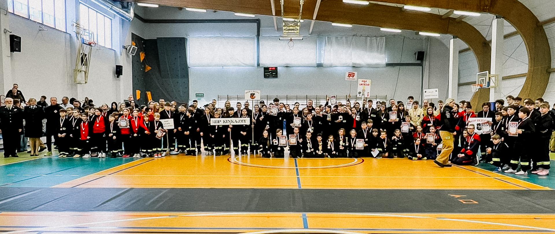 Szerokie ujęcie całej hali sportowej wypełnionej uczestnikami zawodów. Setki dzieci w mundurach i dresach stoją wzdłuż linii boiska, tworząc imponujący widok zakończenia imprezy.