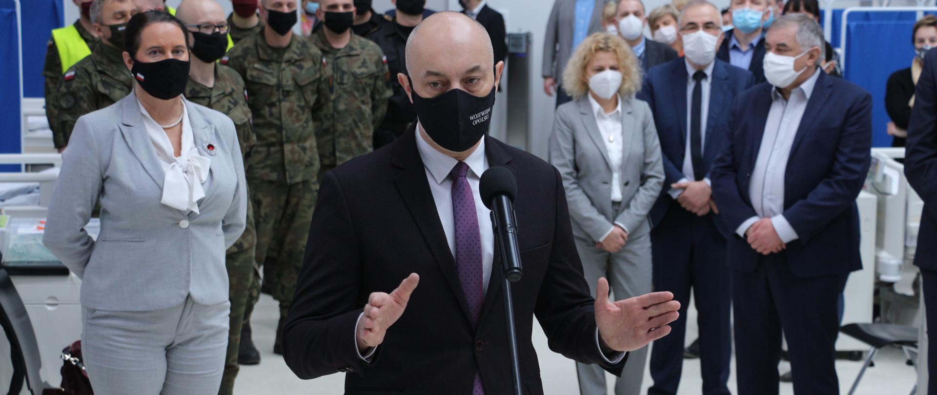 Konferencja prasowa wojewody opolskiego - podsumowanie prac nad budową szpitala tymczasowego w Opolu. Na zdjęciu wojewoda opolski przemawiający przez mikrofon na statywie do zgromadzonych osób. W tle pozostali uczestnicy konferencji oraz łóżka medyczne. 