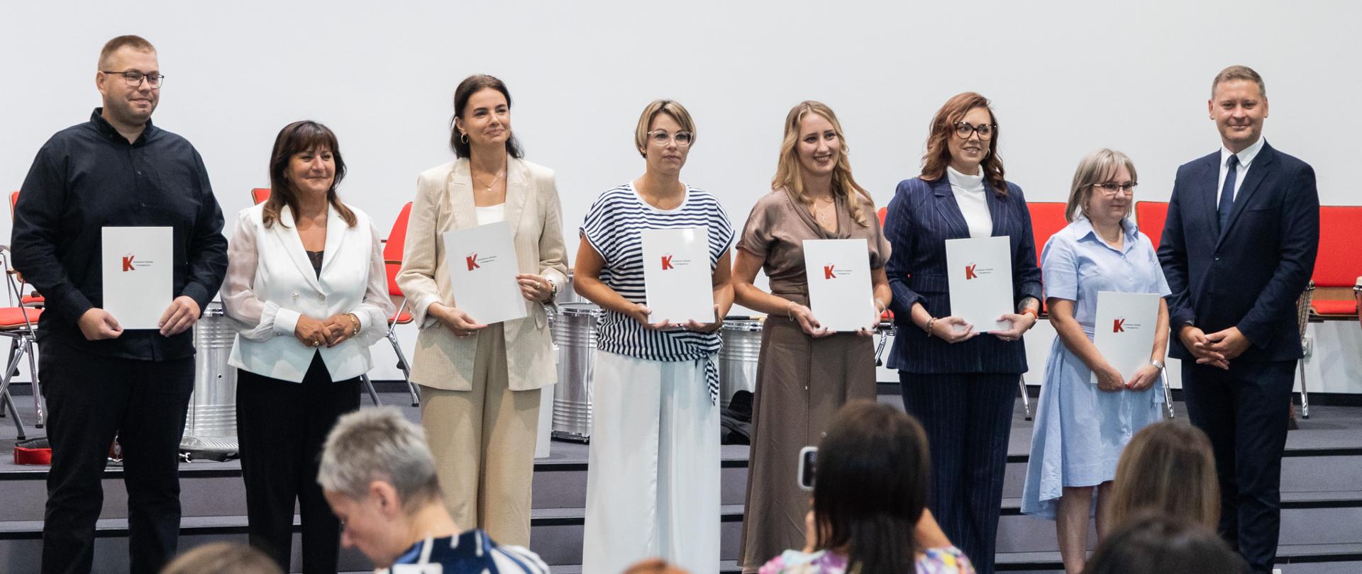 Grupa siedmiu osób trzymających białe teczki z dyplomami stoi w szeregu na scenie. Po prawej stronie stoi wicewojewoda kujawsko-pomorski Michał Koniuch w ciemnym garniturze. Z przodu widać nieostre głowy publiczności obserwującej wręczenie wyróżnień.