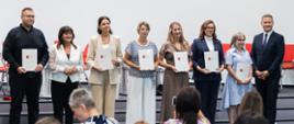 Grupa siedmiu osób trzymających białe teczki z dyplomami stoi w szeregu na scenie. Po prawej stronie stoi wicewojewoda kujawsko-pomorski Michał Koniuch w ciemnym garniturze. Z przodu widać nieostre głowy publiczności obserwującej wręczenie wyróżnień.