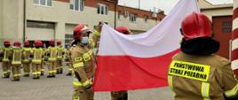 Strażacy stoją w dwóch rzędach na przeciwko siebie, uroczyście podnosząc flagę Rzeczypospolitej Polskiej na maszt.