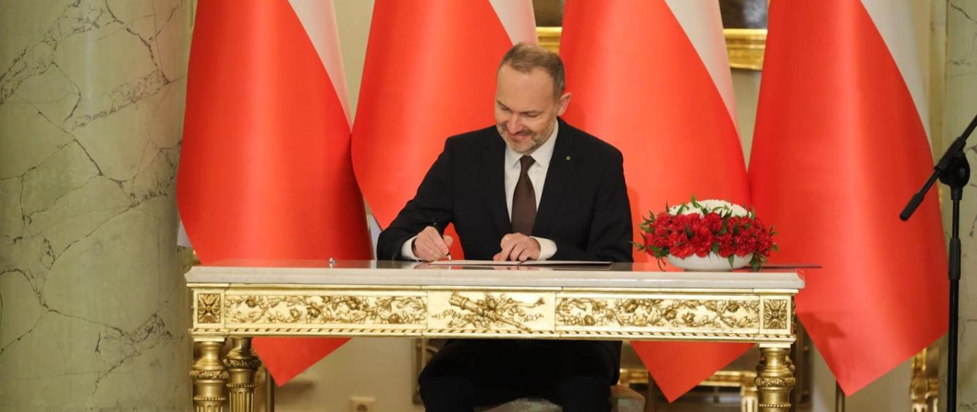 Na zdjęciu widać ministra Krzysztofa Hetmana, który podpisuje akt powołania na ministra. Minister siedzi przy stole na którym po prawej stronie stoją biało czerwone kwiaty. W tle flagi Polski.